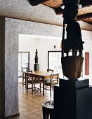 African sculpture on pedestal in front of wide open doorway with view of dining area beyond in modern house with wood-beamed ceiling