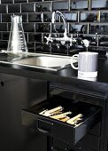 Detail of black kitchen counter with vintage-look sink and open cutlery drawer in base unit