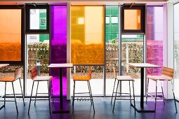 Tall tables and bar stools in front of glass facade with coloured glass panels