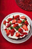 A cream cake with dark base topped with strawberries on a table with a red cloth