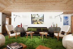 Sessel und Beistelltische im 50er Jahre Stil auf grünem Teppichboden vor Wand mit moderner Wandleuchte im Wohnraum mit Holzdecke