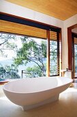 White, designer bathtub on stone floor in front of glass wall with view of sea and trees
