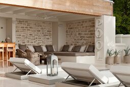 Peaceful, roofed seating area with upholstered platform and many scatter cushions next to pool; modern sun loungers and lantern in foreground