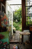 Stack of painting and vintage stool in studio with open glass door and view of garden