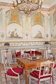 Chairs painted pale grey with red seat cushions around antique table in grand salon with murals on walls