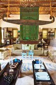 Wooden object hanging from ceiling above wooden table and sofa set in lounge area in front of green, floor-to-ceiling wooden panel in open-plan interior