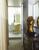 Messingfarbener Torso auf Kommode im Gang mit offener Tür und Blick auf Stuhl vor Waschtisch mit Spiegel