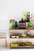 Bird cage next to vases and bowls of various sizes on half-height shelving next to partially visible chairs below pendant lamp against white wall