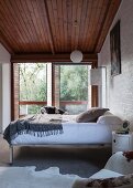 Bed with chrome metal frame against wall in front of terrace doors in plain bedroom with wooden ceiling