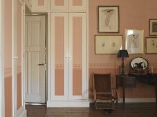 Mit Kleiderschränken umbaute Zimmertür eines stilvollen Schlafraumes; Tönung von Wand und Schranktüren in kalkigem Rose mit umlaufender Bordüre
