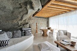 Curved bench with black and white patterned cushions against rock wall and white upholstered furniture in front of glass wall with transparent curtain