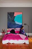 Doppelbett mit pinkfarbenem Bezug im Batiklook vor grossformatigem Farbgemälde; Frau sitzt auf Bett