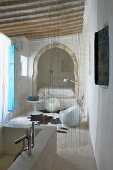 Bad Ensuite mit Natursteinwanne; Schnurvorhang als Abgrenzung zu Sitzplatz mit Designermöbeln und Bett in orientalischem Alkoven