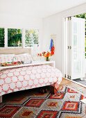 Double bed with overbed table in sunny room with open folding door leading to garden terrace; ethnic-style woven rug on floor