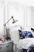 Nostalgic double bed with ruffled lace pillows against white wall with Baroque ornamentation; chromed, angle-poise lamp on bedside table