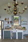 Messing-Kronleuchter in weisser Küche mit grauen Schranktüren und vergittertem Fenster