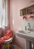 Pedestal washbasin with tiled splashback below mirror hung on pink wall next to wire mesh chair in corner of room