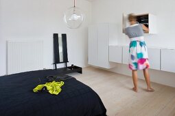 Frau vor weissen Wand- Hängeschränken in minimalistischem Schlafzimmer und gelbgrünes Kleid auf schwarzer Tagesdecke