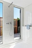Minimalistisches, grau getöntes Bad mit Designer Duscharmatur unter Kopfbrause an Wand und Blick durch raumhohe Fenster auf die Terrasse