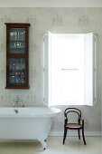 Traditional bathroom with glass-fronted wall cabinet above retro bathtub and classic bentwood chair; window with interior shutters