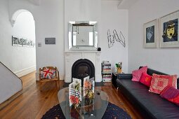 Kidney-shaped glass coffee table and black leather couch in traditional, minimalist interior with open fireplace next to arched doorway leading to narrow hallway