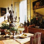 Rustic dining area with magnificent candelabra and fruit and wine bottles on sideboard; still-life paintings and stuffed pheasant on walls