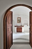 Open double doors of dark wood in arched doorway and view into minimalist bathroom with washstand flanked by niches