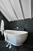Freistehende Badewanne in modernem Bad mit Schieferplatten auf Wand und Boden unter weiss gestrichener Holzdecke