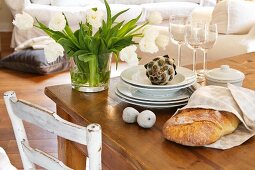A bunch of white tulips in a glass vase next to a stack of plates, some wine glasses, decorative figs and a decorative artichoke