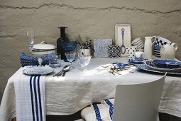 White and blue crockery and glasses on white tablecloth on table against roughly plastered wall