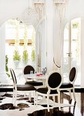 Postmodern dining area - white neo-rococo chairs with black upholstery in front of arched French windows with view onto terrace