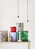 Assorted wallpapered lamp shades on a white, wood table under a hanging light with wire frames