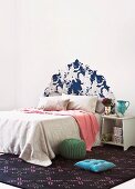 Doppelbett mit Tagesdecken vor Wand mit tapeziertem Kopfteil in barockem Stil und schlichtes Nachttischchen auf gemustertem Teppich mit Bodenkissen und Pouf