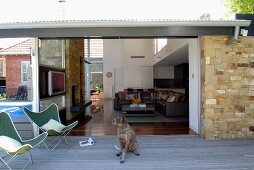 Blick von Holzterrasse mit Fledermaussesseln und Hund in den hohen Wohnraum eines zeitgenössischen Hauses mit viel Glas und Naturstein