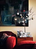Small tree decorated with Christmas decorations and presents in front of dark painting in living room