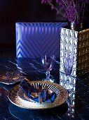 Place setting with gold-rimmed plate and shiny silver vase on dining table in front of chair with elegant upholstered backrest