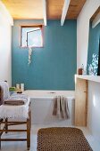 Einfaches Bad mit Badewanne an hellblauer Wand und kleines Fenster, im Vordergrund Stuhl mit Handtüchern und Sisalläufer