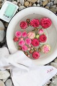 Schwimmende Rosenblüten in alter Emailleschüssel mit weißem Leinenhandtuch und Seifenschale auf einer Kiesfläche arrangiert