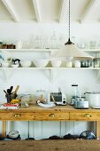 Simple pendant lamp in front of kitchen utensils and appliances on rustic wooden sideboard below bowls and glasses on wall-mounted shelves