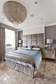Lavish, silver double bed with tall, velvet-covered headboard below magnificent, modern chandelier