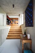 Vintage console table and modern artwork on concrete wall; pale wooden stairs and large bookcase in background