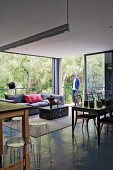 Breakfast bar and plexiglas stools (Ghost), dining area with fifties chairs and sofa and trunk table in open-plan interior with glass walls; man on terrace in background