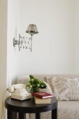 Vintage telephone on round, black side table in front of pale sofa against wall and retro telescopic scone lamp