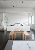 Tisch und Bänke aus hellem Holz auf grauem Estrichboden vor weiss gehaltenem Schlafbereich in loftartigem Raum