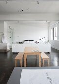 Pale wooden table and benches on grey concrete floor in front of white sleeping area in loft-style interior