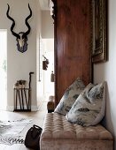 Scatter cushions on ottoman next to antique cupboard; hunting trophy on wall in background