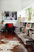 Kuhfell und Bücherstapel auf Boden vor halbhohem Regal unter Fensterband, im Hintergrund Sessel & grossformatiges Foto an Wand