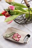 Handy in handbestickter Tasche neben Drahtkorb mit Frühlingsblumen