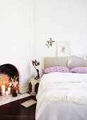 Double bed with upholstered headboard next to open fireplace in white-painted brick wall