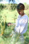 Young woman in romantic summer clothing with wicker basket standing in front of pine branches